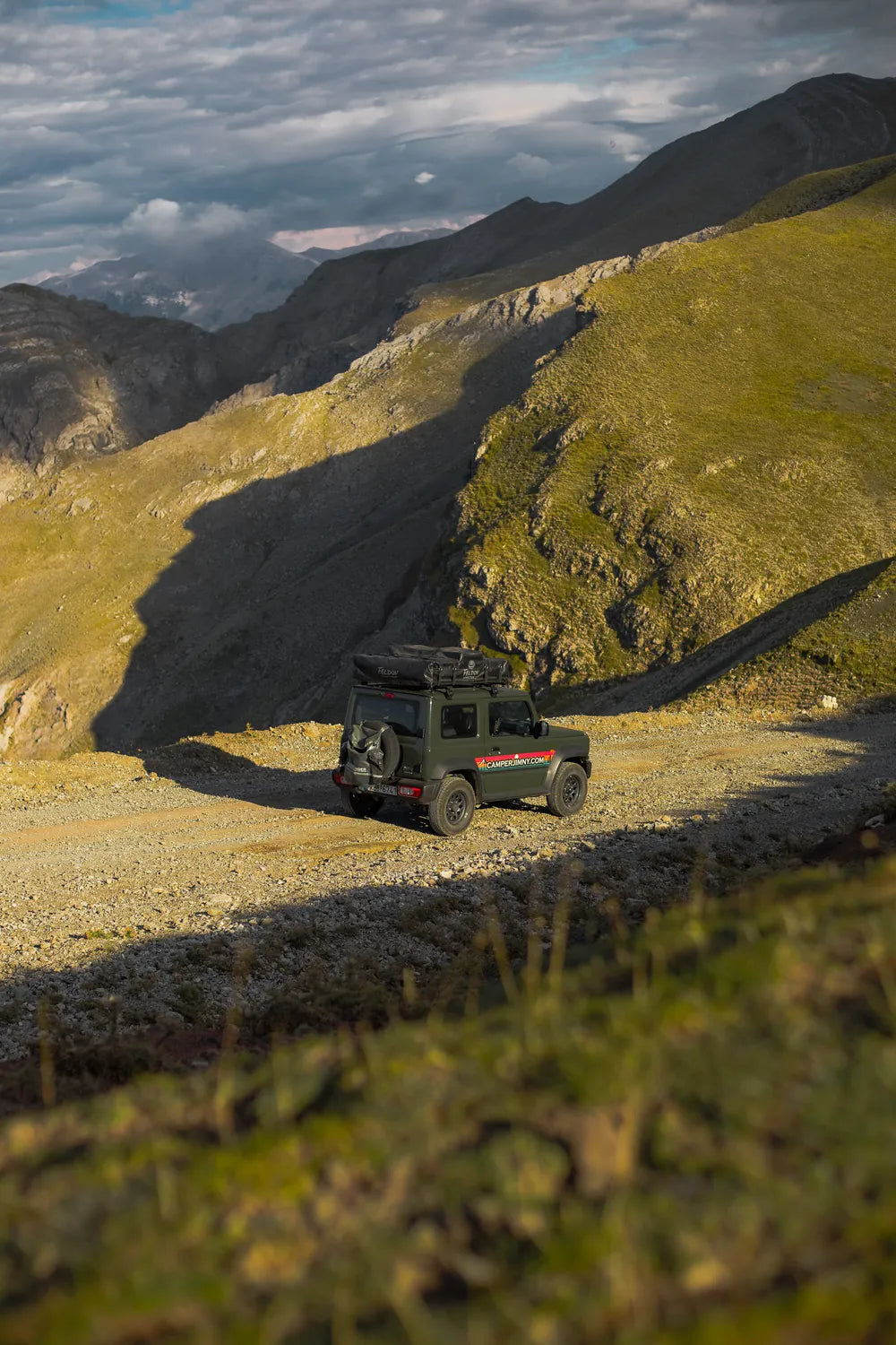 Green Suzuki Jimny driving a mountain road below green slopes on an overland trip in Greece