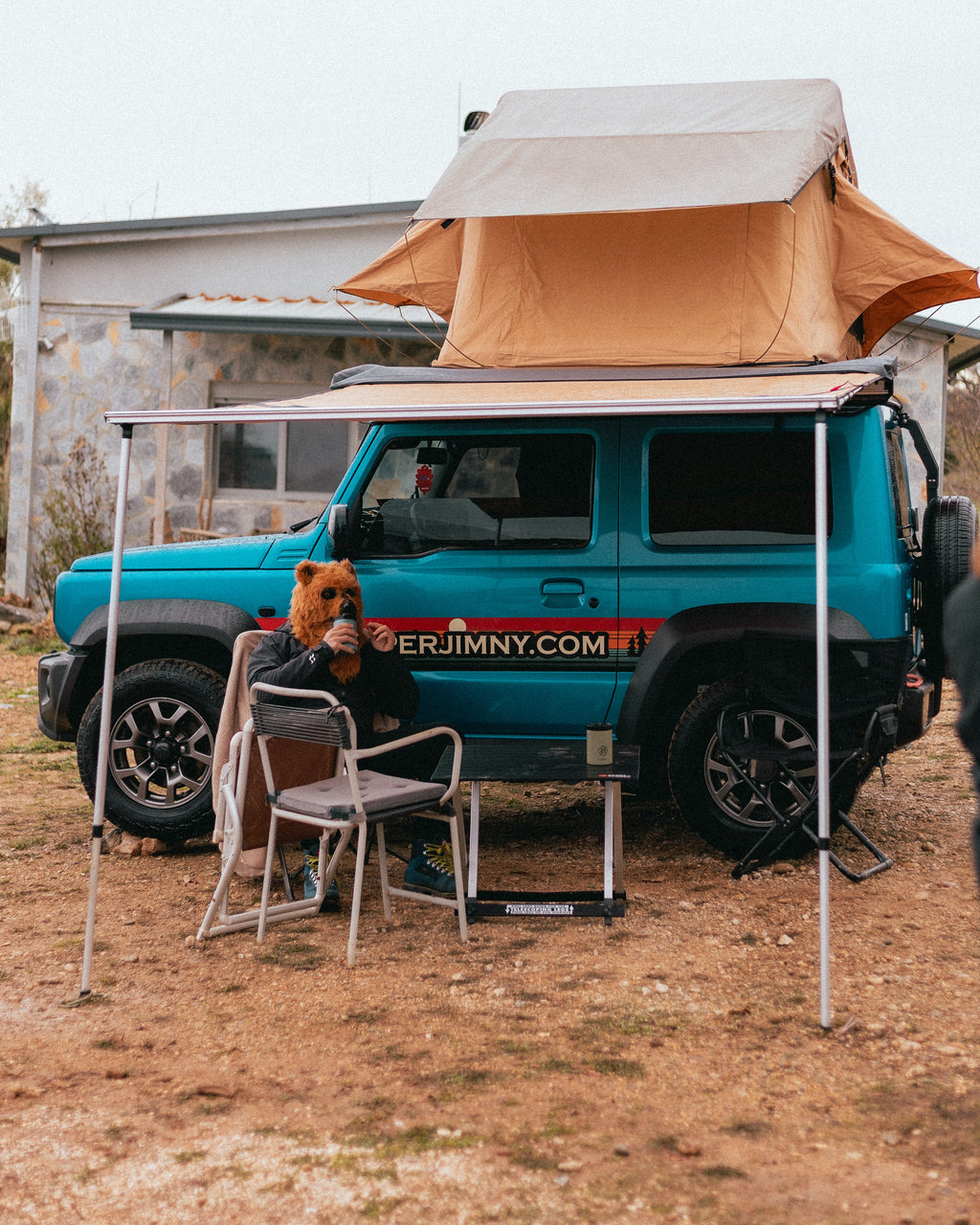 A Suzuki Jimny with a roof top tent and side awning set up at camp with camping chairs