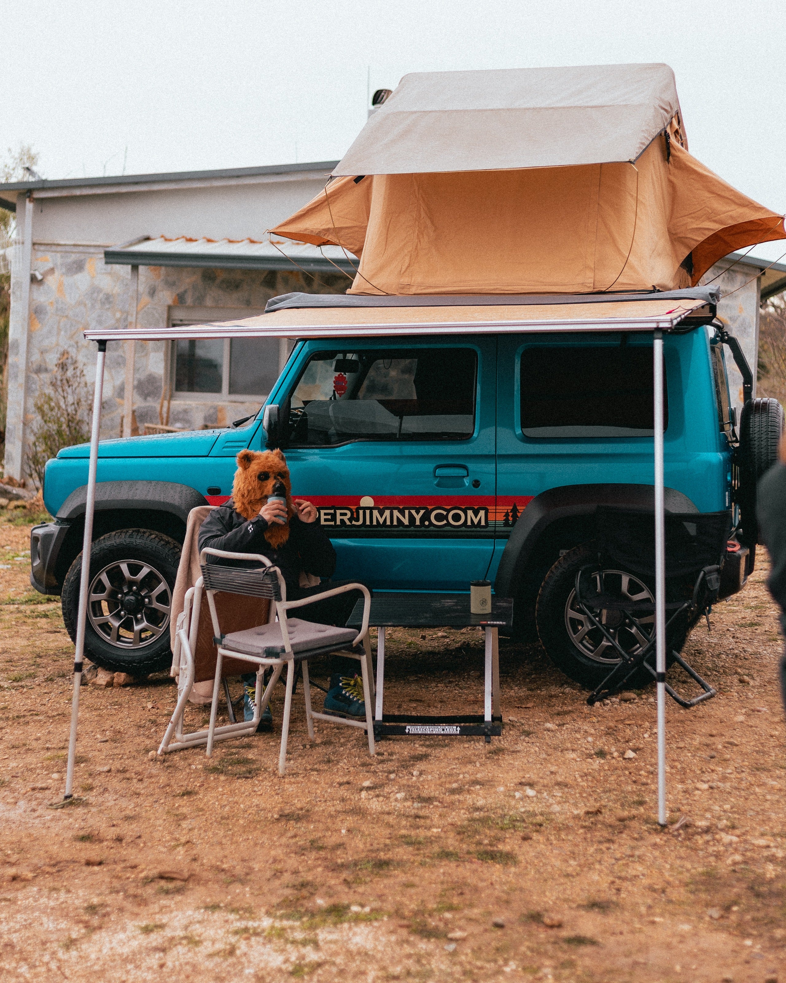A Suzuki Jimny with a roof top tent and side awning set up at camp with camping chairs