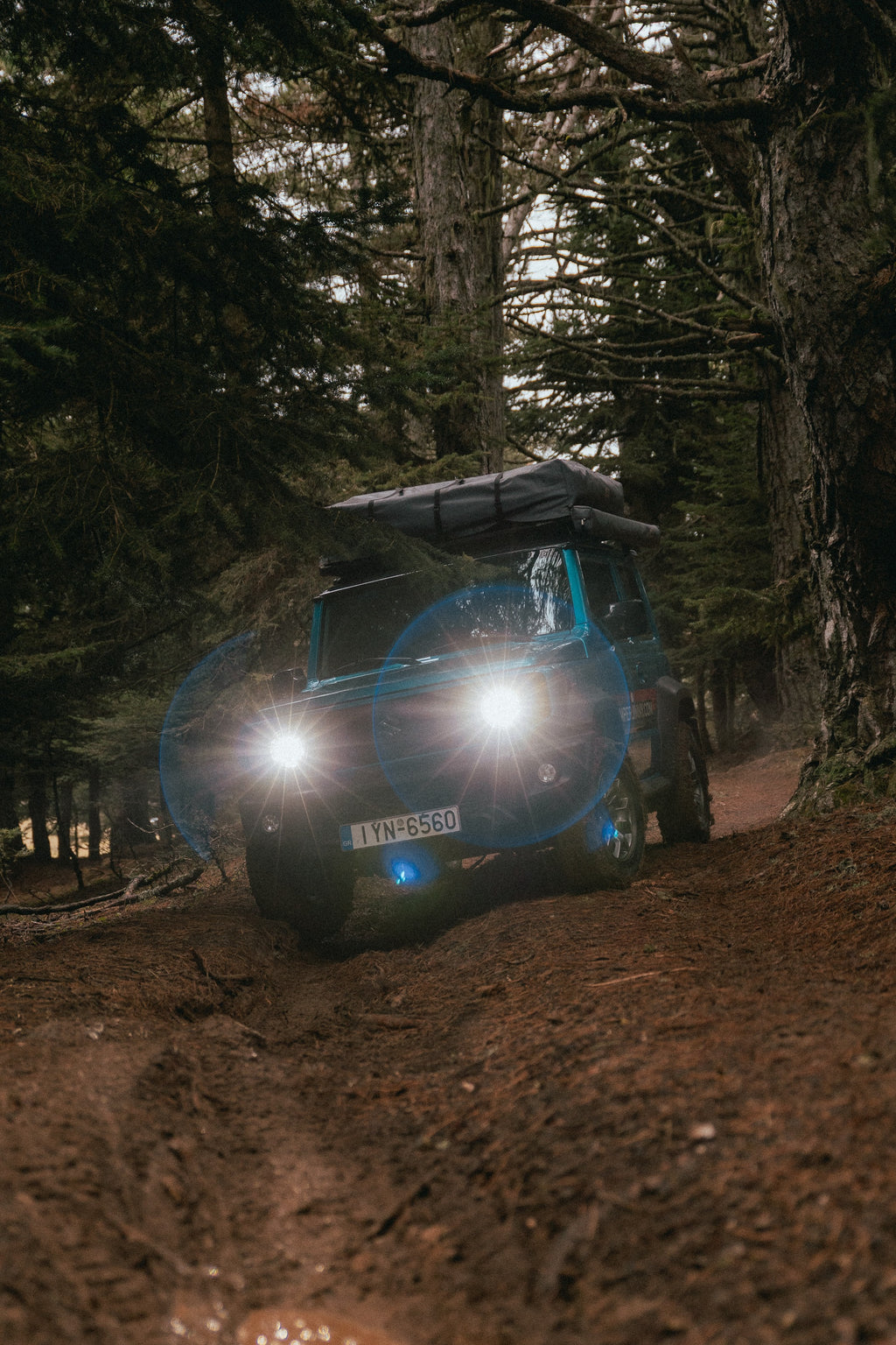 A Suzuki Jimny with headlights on driving a wet forest track through fir trees
