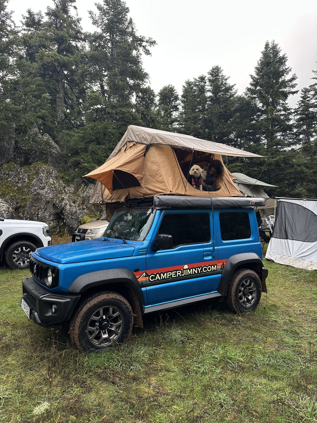 A blue Suzuki Jimny with its roof top tent open at a forest camp